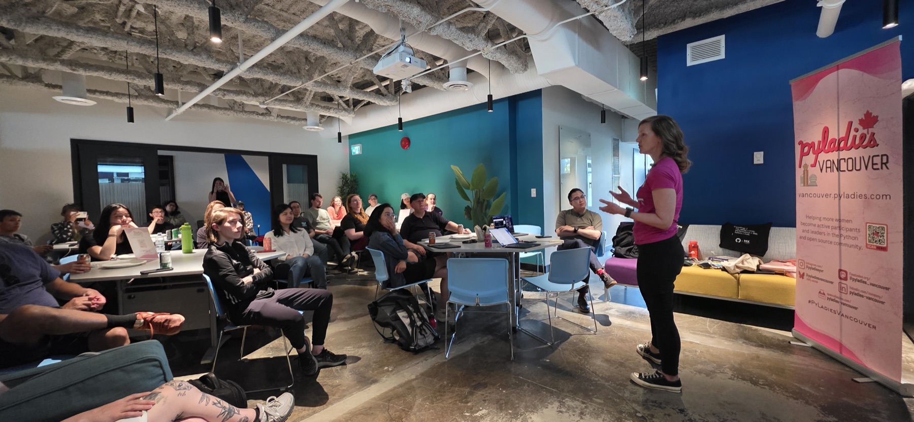 A full room of attendees at the July meetup, seated around tables and listening to a talk, with the PyLadies Vancouver banner at the front
